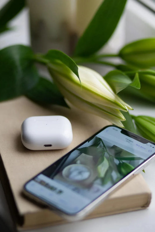 An AirPods case and a smartphone on top of a book near a  flower.