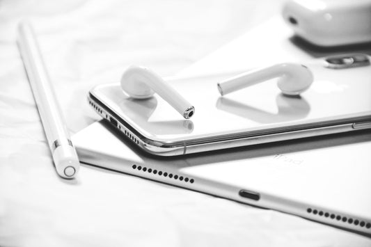 An iPad, iPhone , stylus, AirPods case, and AirPods on white surface.