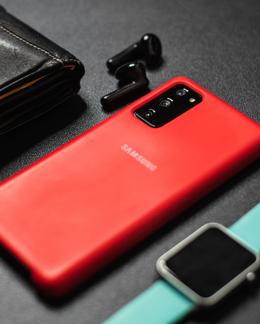 A red Samsung phone on a table next to  earphones, wallet, and a smartwatch.